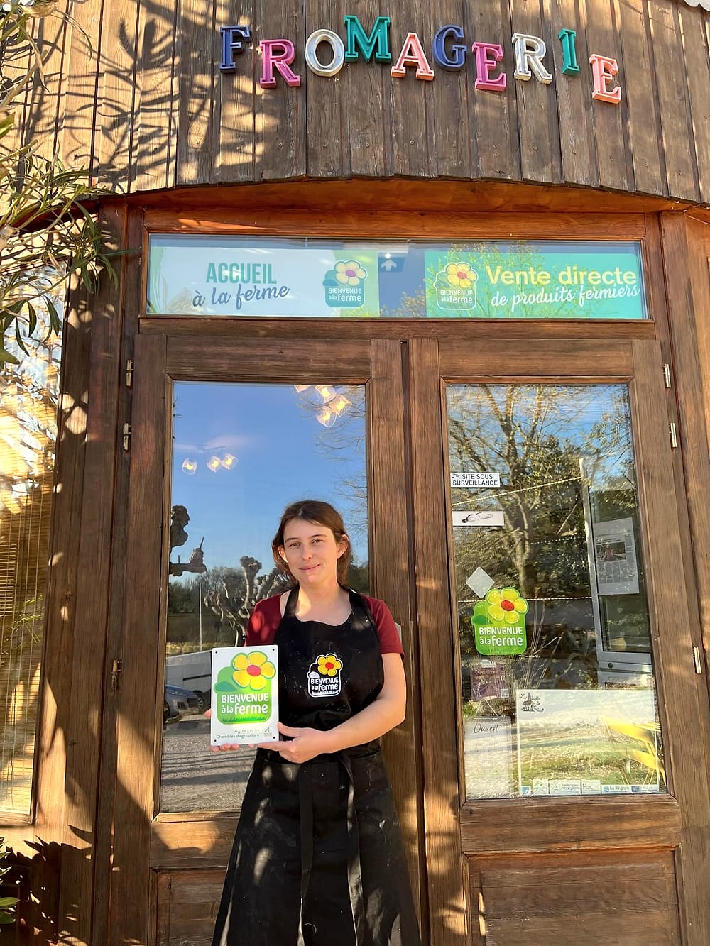 Laura Larchevêque, en tablier, devant la porte en bois du magasin de la fromagerie, tient la plaque du label Bienvenue à la ferme