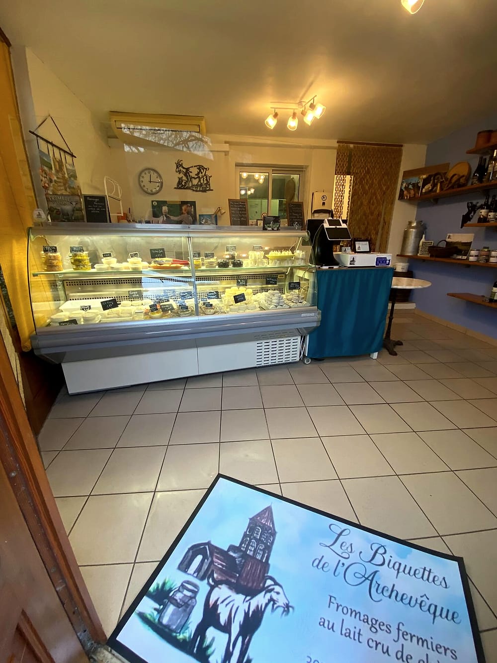 L'intérieur du magasin : la vitrine à fromages, le comptoir et les étagères de l'épicerie, avec le tapis au logo de la ferme à l'entrée
